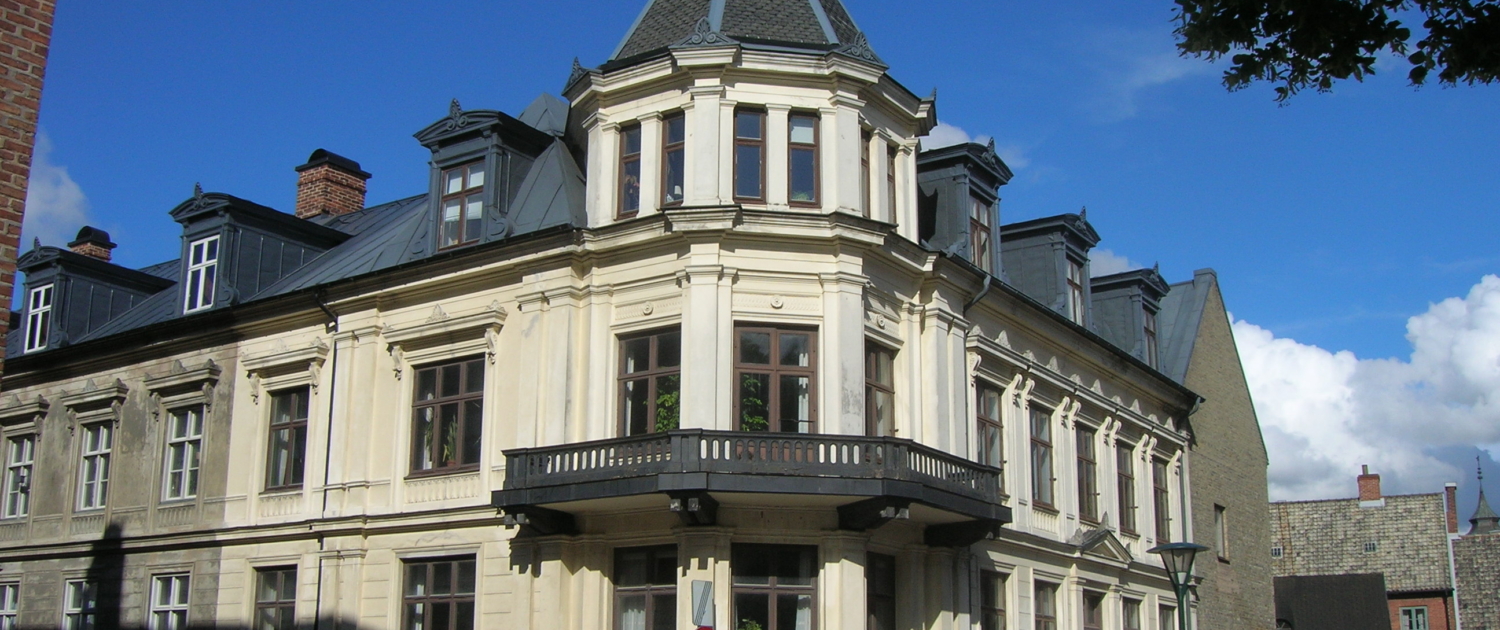 Historisk hörnbyggnad i ljus puts med mörkt plåttak och ett runt torn med spetsig kupol; en liten balkong löper längs fasaden ovanför entrén, och huset ligger i stadsmiljö med kullerstensgata, parkerade bilar och blå himmel med spridda moln.